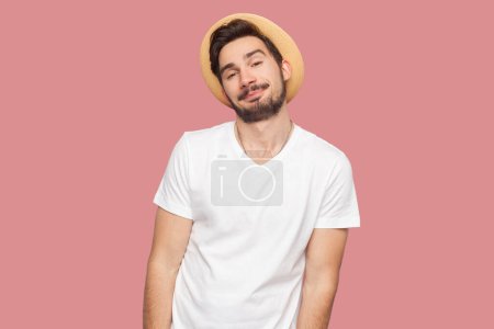 Téléchargez les photos : Portrait de cool homme barbu souriant et confiant en t-shirt blanc et chapeau debout regardant la caméra, ayant expression satisfaite. Studio intérieur tourné isolé sur fond rose. - en image libre de droit