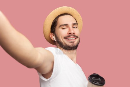 Photo for Portrait of handsome cheerful joyful bearded man in white T-shirt and hat standing drinking coffee, looking smiling at camera, POV. Indoor studio shot isolated on pink background. - Royalty Free Image