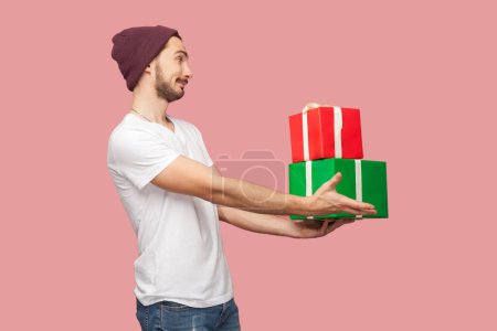 Photo for Side view portrait of smiling happy bearded man in white T-shirt and beany hat standing giving gift to his friend congratulating with birthday. Indoor studio shot isolated on pink background. - Royalty Free Image
