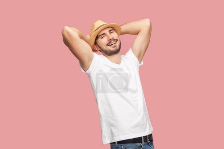 Foto de Retrato de hombre hipster barbudo guapo relajado satisfecho en camiseta blanca y sombrero casual de pie con las manos levantadas, mirando a la cámara. Estudio interior plano aislado sobre fondo rosa. - Imagen libre de derechos