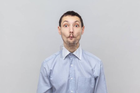 Photo for Portrait of funny childish young adult man standing looking at camera, playing with kids, making fish lips, wearing light blue shirt. Indoor studio shot isolated on gray background. - Royalty Free Image