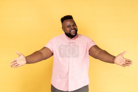 Téléchargez les photos : Portrait d'homme barbu heureux et généreux portant une chemise rose debout avec les mains levées et regardant la caméra, accueillant ou donnant smth. Studio intérieur tourné isolé sur fond jaune. - en image libre de droit