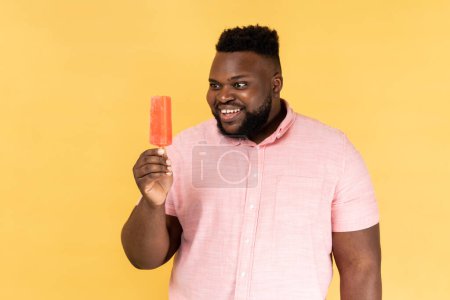 Téléchargez les photos : Portrait d'un homme souriant portant une chemise rose tenant de la crème glacée douce veut essayer de délicieux desserts de confiserie dans ses mains. Studio intérieur tourné isolé sur fond jaune. - en image libre de droit