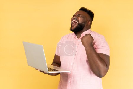 Téléchargez les photos : Portrait d'un homme extrêmement heureux portant une chemise rose criant de joie et tenant un ordinateur portable, se réjouissant de la victoire, paris en ligne. Studio intérieur tourné isolé sur fond jaune. - en image libre de droit