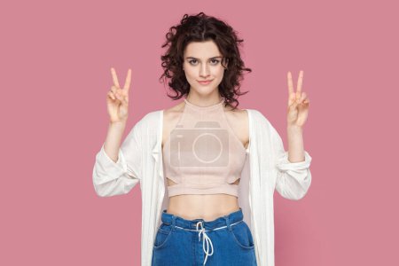 Photo for Portrait of attractive beautiful young adult woman with curly hair wearing casual style outfit standing looking at camera, showing v sign. Indoor studio shot isolated on pink background. - Royalty Free Image