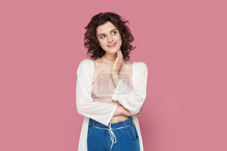 Photo for Portrait of friendly pleased smiling woman with curly hair wearing casual style outfit, looking away, dreaming about something pleasant. Indoor studio shot isolated on pink background. - Royalty Free Image
