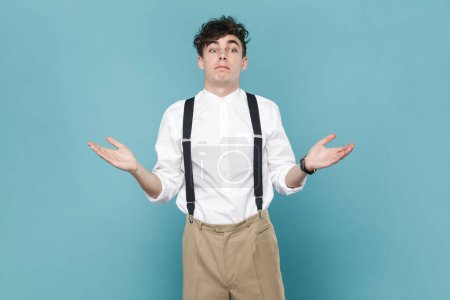 Photo for Portrait of puzzled confused man wearing white shirt and suspender, shrugging shoulders, looking away, raised his arms, has uncertain expression. Indoor studio shot isolated on blue background. - Royalty Free Image