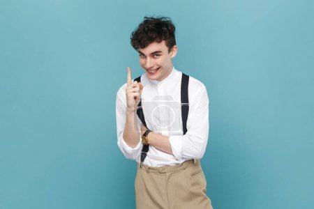 Photo for Portrait of clever smart smiling excited man wearing white shirt and suspender raising finger up, having idea for new business project. Indoor studio shot isolated on blue background. - Royalty Free Image
