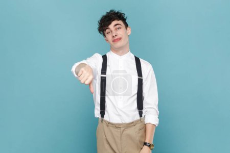 Photo for Portrait of disappointed sad upset handsome man wearing white shirt and suspender showing thumb down, demonstrates dislike gesture. Indoor studio shot isolated on blue background. - Royalty Free Image