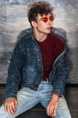 Photo for Portrait of trendy stylish attractive fashion model man in bright red sunglasses and denim casual style sitting on floor near metallic wall and looking away. Indoor studio shot. - Royalty Free Image