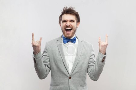 Photo for Portrait of crazy funny excited handsome bearded man standing with rock and roll gesture, screaming with excitement, wearing grey suit and blue bow tie. Indoor studio shot isolated on gray background. - Royalty Free Image