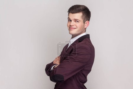 Photo for Side view portrait of smiling optimistic young man standing looking ahead, expressing happiness, keeps arms crossed, wearing violet suit and white shirt. Indoor studio shot isolated on grey background - Royalty Free Image