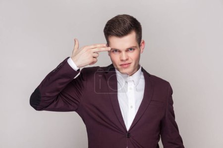 Photo for Kill me please. Depressed man committing suicide with finger gun gesture, shooting himself, being hysterical, wearing violet suit and white shirt. Indoor studio shot isolated on grey background. - Royalty Free Image