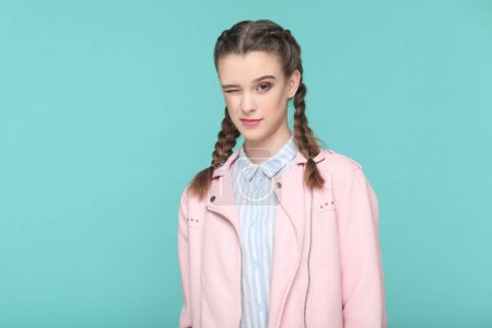 Photo for Portrait of playful cheerful teenager girl with braids wearing pink jacket standing looking at camera, winking, flirting with boyfriend. Indoor studio shot isolated on green background. - Royalty Free Image
