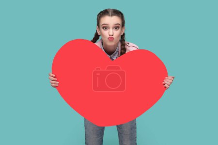 Photo for Portrait of optimistic romantic lovely teenager girl with braids wearing pink jacket holding big red heart, sending air kissing. Indoor studio shot isolated on green background. - Royalty Free Image