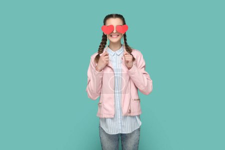 Téléchargez les photos : Portrait d'adolescente romantique positive avec des tresses veste rose debout couvrant ses yeux avec deux coeurs rouges sur le bâton. Studio intérieur tourné isolé sur fond vert. - en image libre de droit