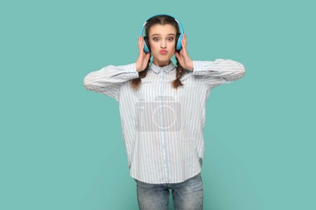 Photo for Portrait of funny teenager girl with braids wearing shirt standing looking at camera with pout lips, listening music, holding hands on headphones. Indoor studio shot isolated on green background. - Royalty Free Image