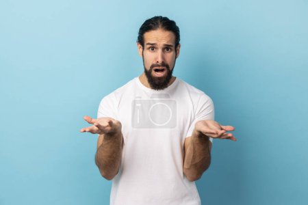 Photo for What do you want? Confused man with beard wearing white T-shirt standing with raised hands and surprised indignant expression, asking what reason. Indoor studio shot isolated on blue background. - Royalty Free Image