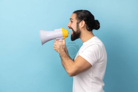 Photo for Side view portrait of handsome man bodybuilder with beard wearing white T-shirt screaming in megaphone, announcing important information. Indoor studio shot isolated on blue background. - Royalty Free Image