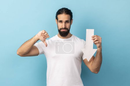 Photo for Portrait of disappointed sad handsome man with beard wearing white T-shirt showing white arrow pointing down, showing thumb down, dislike gesture. Indoor studio shot isolated on blue background. - Royalty Free Image
