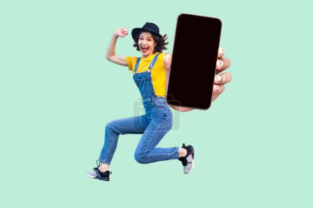Photo for Happy carefree girl in blue denim overalls, yellow shirt and black hat jumping, showing big cell phone with black screen with advertisement area. Indoor studio shot isolated on light green background. - Royalty Free Image