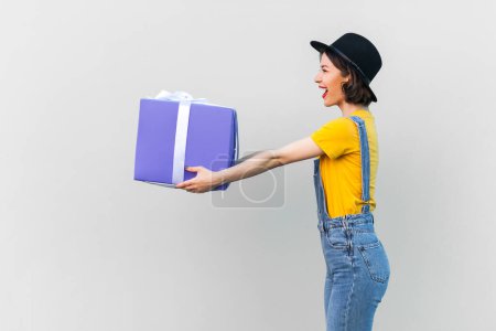 Photo for Side view portrait of amazed hipster woman in blue denim overalls, yellow T-shirt and black hat, giving present box, congratulate with holiday. Indoor studio shot isolated on gray background. - Royalty Free Image