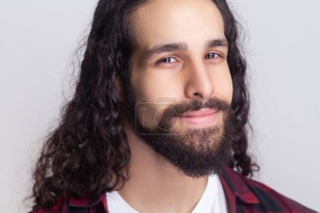 Photo for Closeup portrait of calm handsome bearded man with long curly hair in checkered red shirt looking at camera with smile, expressing positive emotions. Indoor studio shot isolated on gray background. - Royalty Free Image