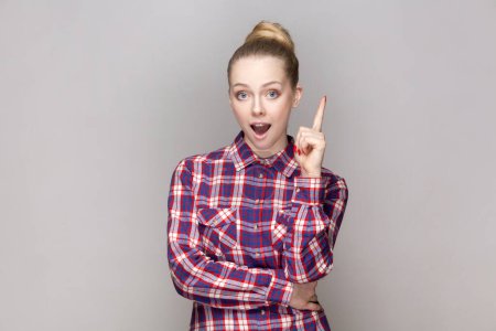 Photo for Portrait of excited smart clever woman with bun hairstyle raised her index finger, having good idea for work project, wearing checkered shirt. Indoor studio shot isolated on gray background. - Royalty Free Image