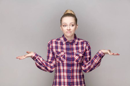 Photo for Portrait of uncertain confused attractive woman with bun hairstyle shrugging shoulders, dosen't know answer, wearing checkered shirt. Indoor studio shot isolated on gray background. - Royalty Free Image