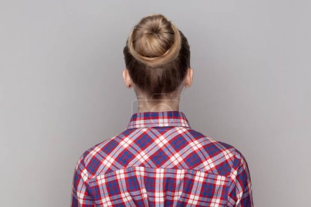 Téléchargez les photos : Vue de dos portrait de femme aux cheveux bruns avec une coiffure de chignon, debout vers l'arrière montrant sa coiffure, portant une chemise à carreaux. Studio intérieur tourné isolé sur fond gris. - en image libre de droit