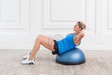 Photo for Portrait of sporty athletic blonde woman with perfect body in black shorts and blue top doing exercises for abdominal muscles on bosu balance trainer, having workout. Indoor studio shot. - Royalty Free Image