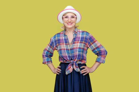 Photo for Cheerful positive optimistic senior woman wearing checkered shirt, hat and eyeglasses standing with hands on hips, looking at camera with happiness. Indoor studio shot isolated on yellow background. - Royalty Free Image