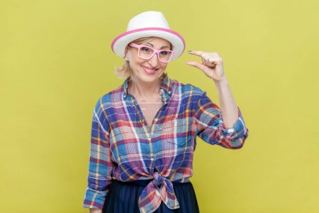 Photo for Portrait of senior woman wearing checkered shirt, hat and eyeglasses shows something very small or tiny, discusses prices in store, smiles happily. Indoor studio shot isolated on yellow background. - Royalty Free Image