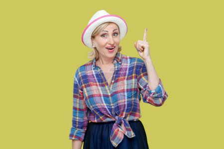 Photo for Portrait of senior woman wearing checkered shirt and hat get new idea, raises fore finger, ready to start work on project, confident in success. Indoor studio shot isolated on yellow background. - Royalty Free Image