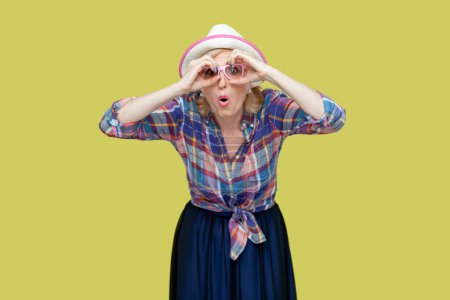 Photo for Senior woman makes binoculars gesture over eyes keeps mouth opened from wonder cannot believe in something awesome concentrated into distance. Indoor studio shot isolated on yellow background. - Royalty Free Image