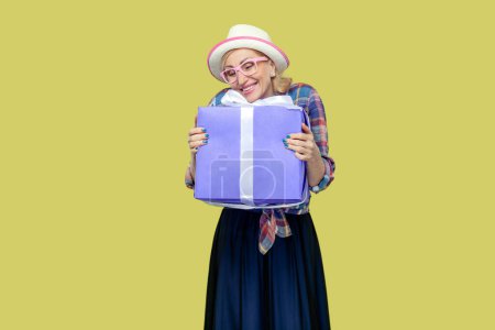 Photo for Portrait of pretty optimistic smiling mature woman wearing checkered shirt, hat and eyeglasses embracing present box, celebrating. Indoor studio shot isolated on yellow background. - Royalty Free Image