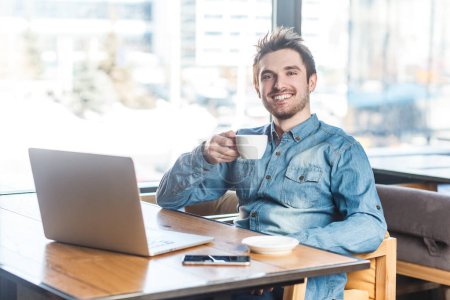 Photo for Portrait of delighted smiling happy young man freelancer in blue jeans shirt working on laptop, having break, drinking coffee. Indoor shot near big window, cafe background. - Royalty Free Image