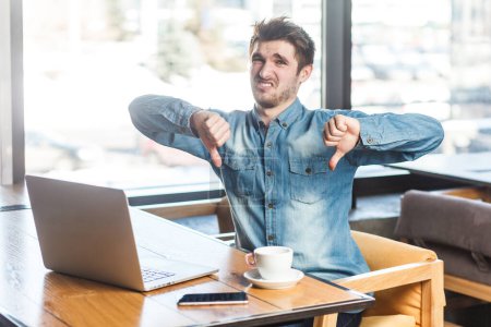 Photo for Portrait of disappointed young man freelancer in blue jeans shirt working on laptop, looking at camera, showing dislike gesture, thumb down. Indoor shot near big window, cafe background. - Royalty Free Image