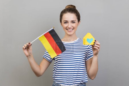 Téléchargez les photos : Portrait de femme satisfaite portant un T-shirt rayé tenant le drapeau allemand et la maison en papier, rêvant d'acheter sa propre maison ou appartement en Allemagne. Studio intérieur tourné isolé sur fond gris. - en image libre de droit