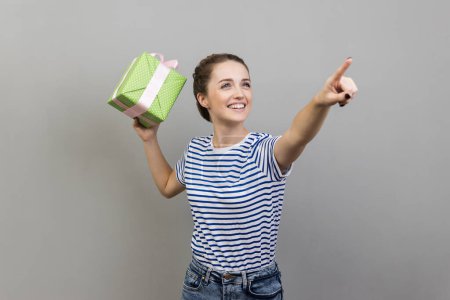 Photo for Portrait of satisfied beautiful woman wearing striped T-shirt throw off your gift box, looking and pointing away and smiling. Indoor studio shot isolated on gray background. - Royalty Free Image