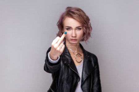 Photo for Portrait of serious rude woman with short hairstyle standing showing middle finger, impolite gesture, looking at camera, wearing black leather jacket. Indoor studio shot isolated on grey background. - Royalty Free Image