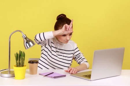 Photo for Lost job, unemployment. Depressed woman sitting at workplace with laptop and showing loser gesture, desperate about unlucky dismissal. Indoor studio studio shot isolated on yellow background. - Royalty Free Image