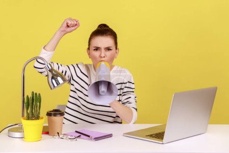 Photo for Portrait of angry woman loudly screaming at megaphone, making announce, protesting, wants to be heard, sitting on workplace. Indoor studio studio shot isolated on yellow background. - Royalty Free Image