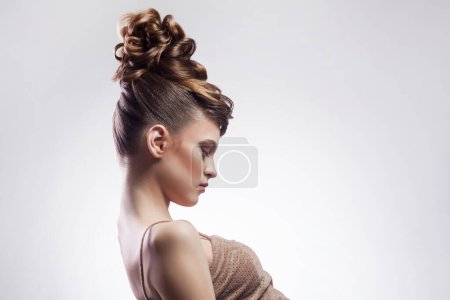 Téléchargez les photos : Portrait de vue latérale de belle jeune femme brune avec coiffure et maquillage élégant, détournant les yeux, démontre sa coiffure à la mode. Studio intérieur tourné isolé sur fond gris. - en image libre de droit