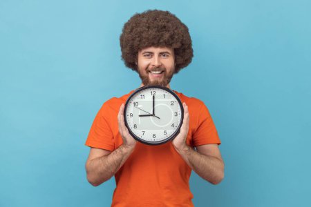 Photo for Portrait of satisfied delighted man with Afro hairstyle wearing orange T-shirt holding in hands big wall clock looking at camera with happy expression. Indoor studio shot isolated on blue background. - Royalty Free Image