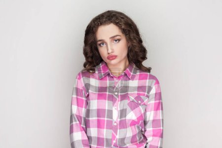 Photo for Portrait of sad lonely woman purses lips, feels bored looks uninterested and tired, listens boring stories from friend, wearing pink checkered shirt. Indoor studio shot isolated on gray background. - Royalty Free Image