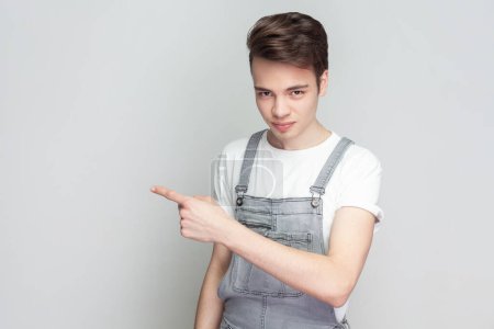 Photo for Get out. Portrait of serious angry young brunette man standing pointing finger, sneak male blaming another people, wearing denim overalls. Indoor studio shot isolated on gray background. - Royalty Free Image