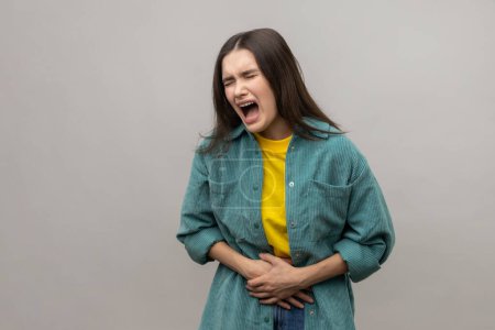 Photo for Stomach ache. Portrait of dark haired woman hunching and holding her belly, suffering period cramps, abdominal pain, wearing casual style jacket. Indoor studio shot isolated on gray background. - Royalty Free Image