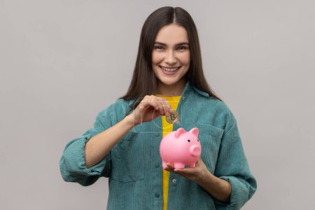 Photo for Portrait of happy smiling dark haired woman investing in bitcoins, putting golden crypto coin into piggy bank, wearing casual style jacket. Indoor studio shot isolated on gray background. - Royalty Free Image