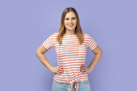 Photo for Portrait of happy satisfied blond woman wearing striped T-shirt standing looking at camera with positive facial expression, keeping hands on hips. Indoor studio shot isolated on purple background. - Royalty Free Image
